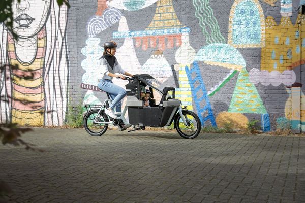 Une femme transporte des enfants dans un vélo cargo sur fond de muraille peinte